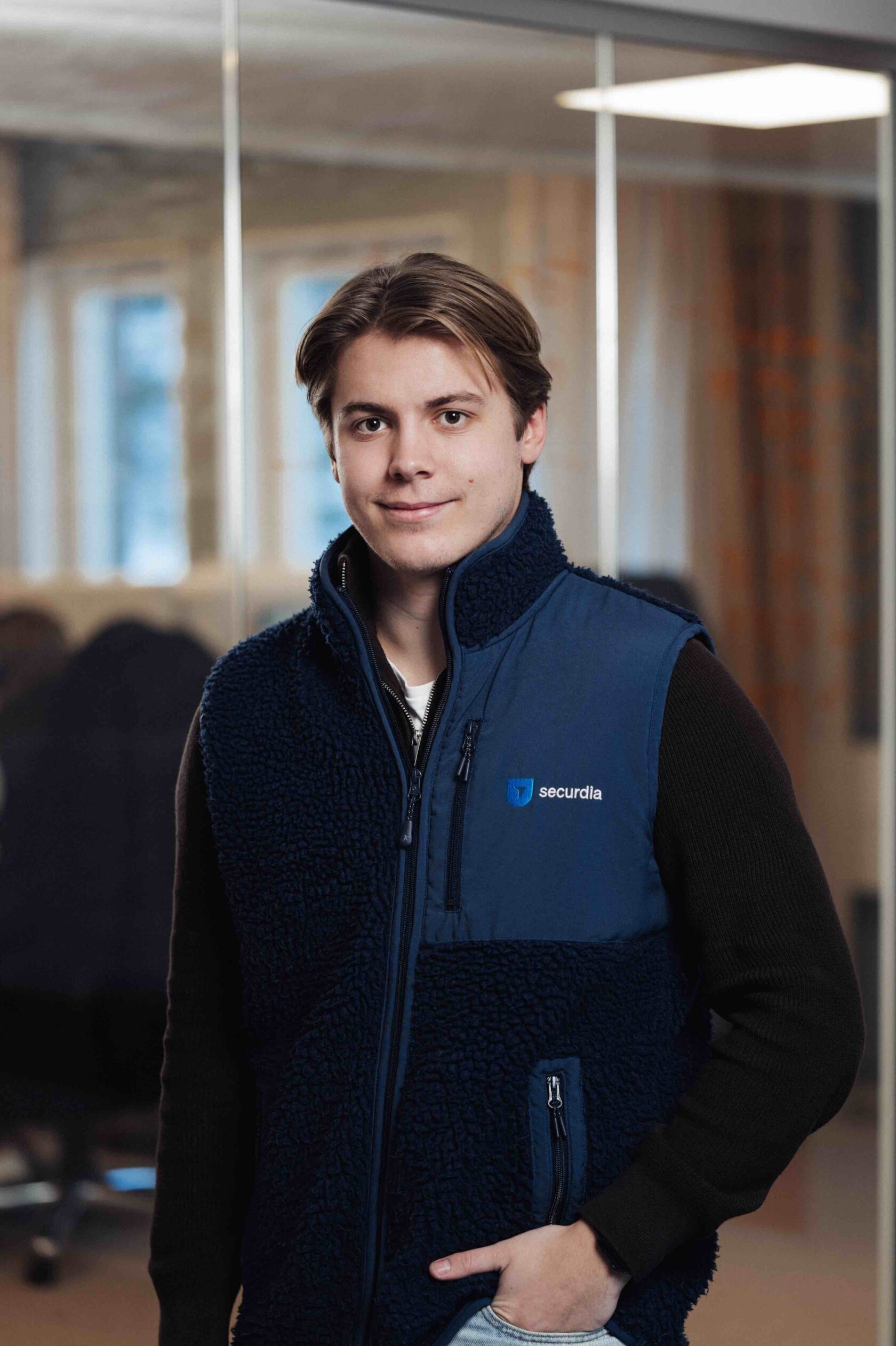 Jonge man in een blauw vest met het logo "securdia" staat binnen en lacht naar de camera.