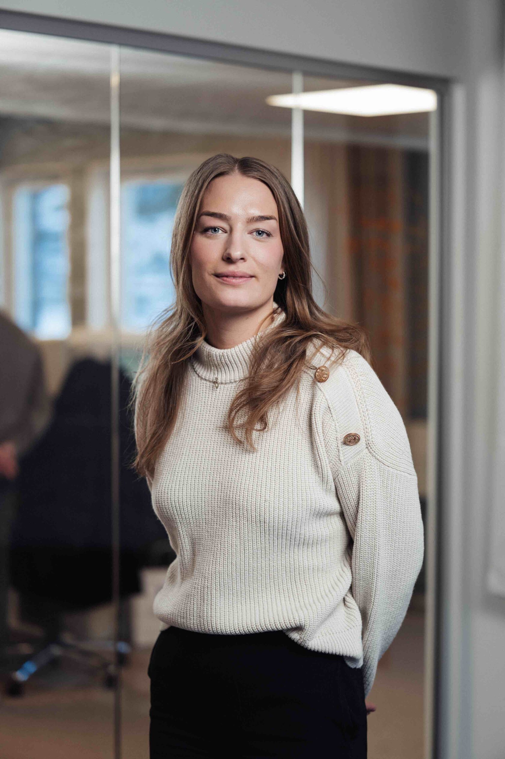 Vrouw met lang bruin haar in een wit gebreide trui staat in een kantoorruimte met glazen wanden op de achtergrond.