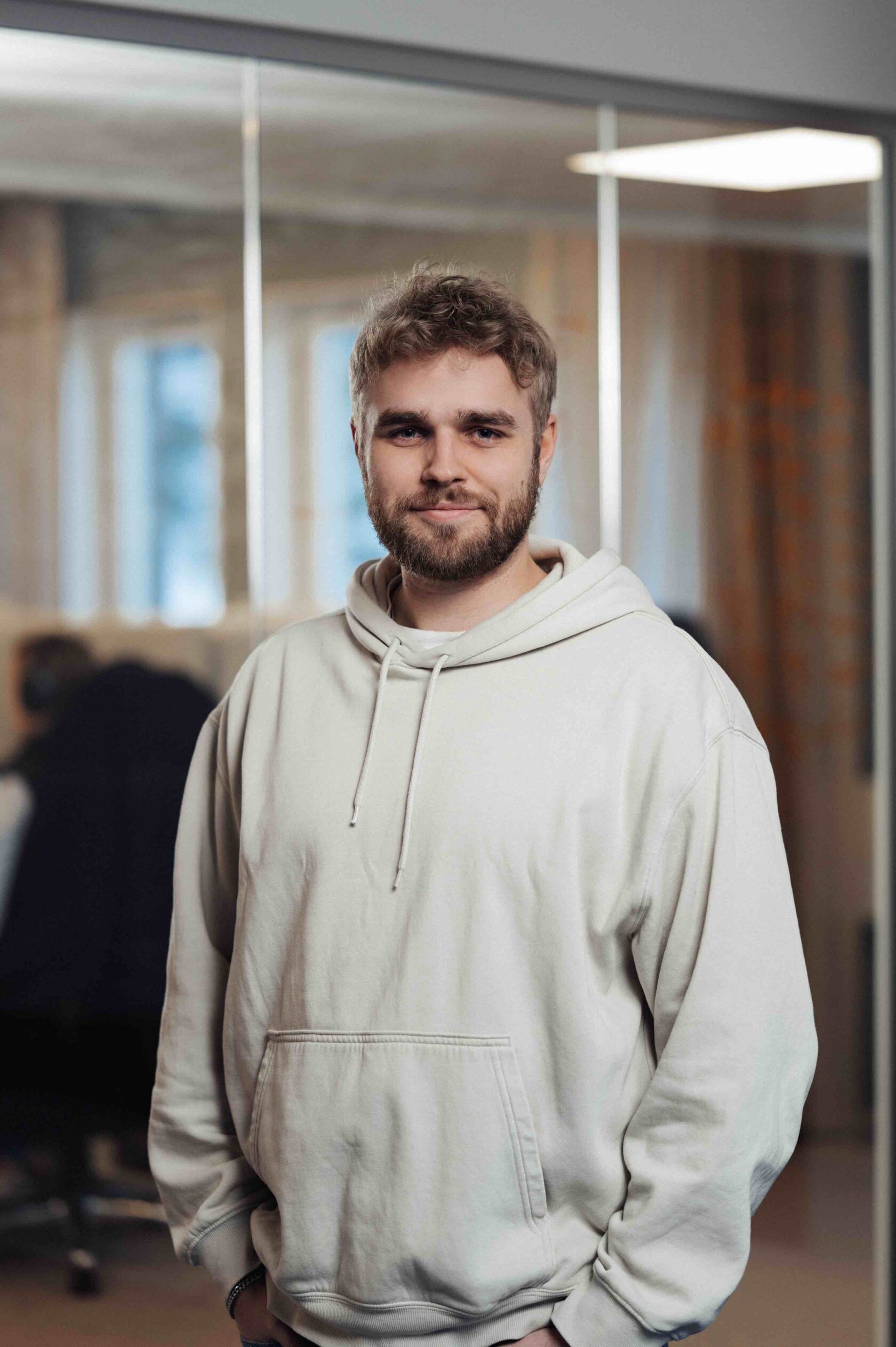 Lachende man met baard in een lichtkleurige hoodie staat binnen met een kantooromgeving op de achtergrond.