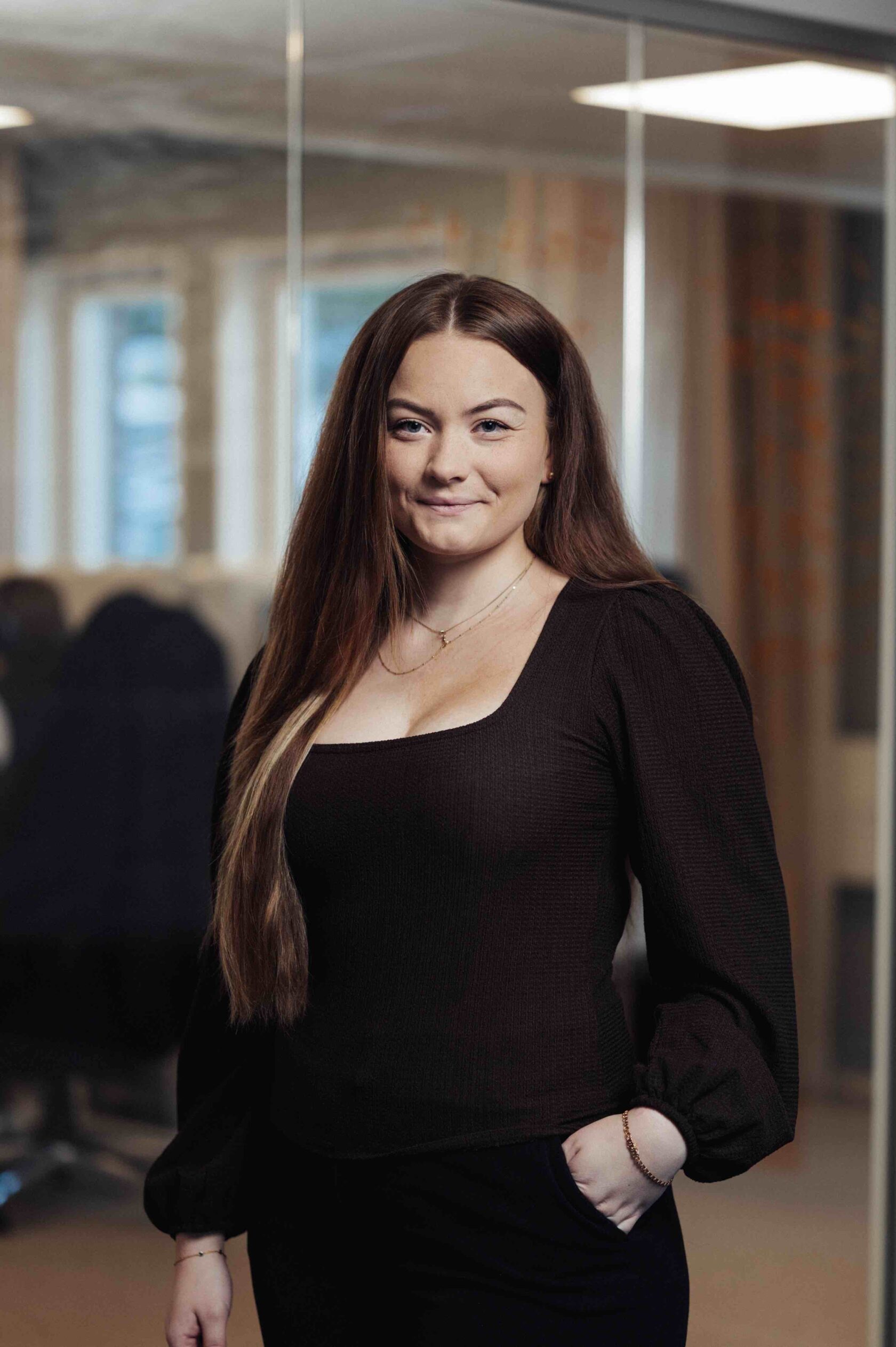 Vrouw met lang bruin haar in een zwarte trui staat in een kantoor en lacht naar de camera.