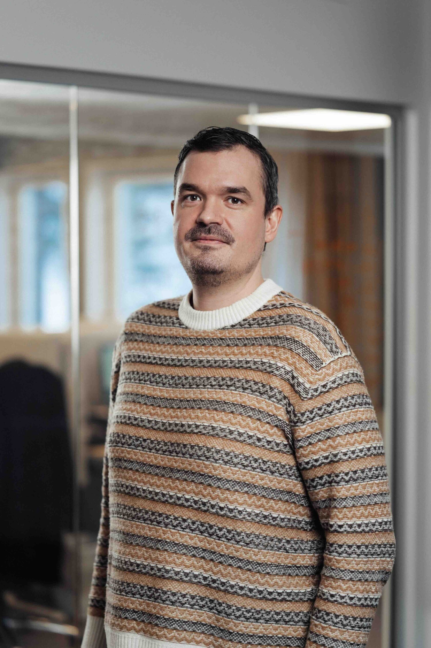 Man in een gebreide trui staat binnen voor een glazen wand met een wazige achtergrond.